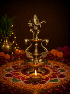 An ornate brass Ganesha idol on a decorative pedestal lamp stand, placed on a lit diya over a red and orange rangoli design, with marigold flowers and warm bokeh lights in the background.