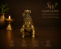 A brass baby figurine of Lord Krishna in a crawling pose, displayed on a dark brown surface with a lit diya and a potted plant in the background. The image features branding for 'Yashrahomes' and feature icons for premium brass, innovative design, artisan craftsmanship, and timeless elegance.