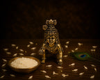 A small metallic brass Lord Krishna figurine kneeling on a dark wooden surface, flanked by a bowl of white rice and a peacock feather, with scattered rice grains around it.