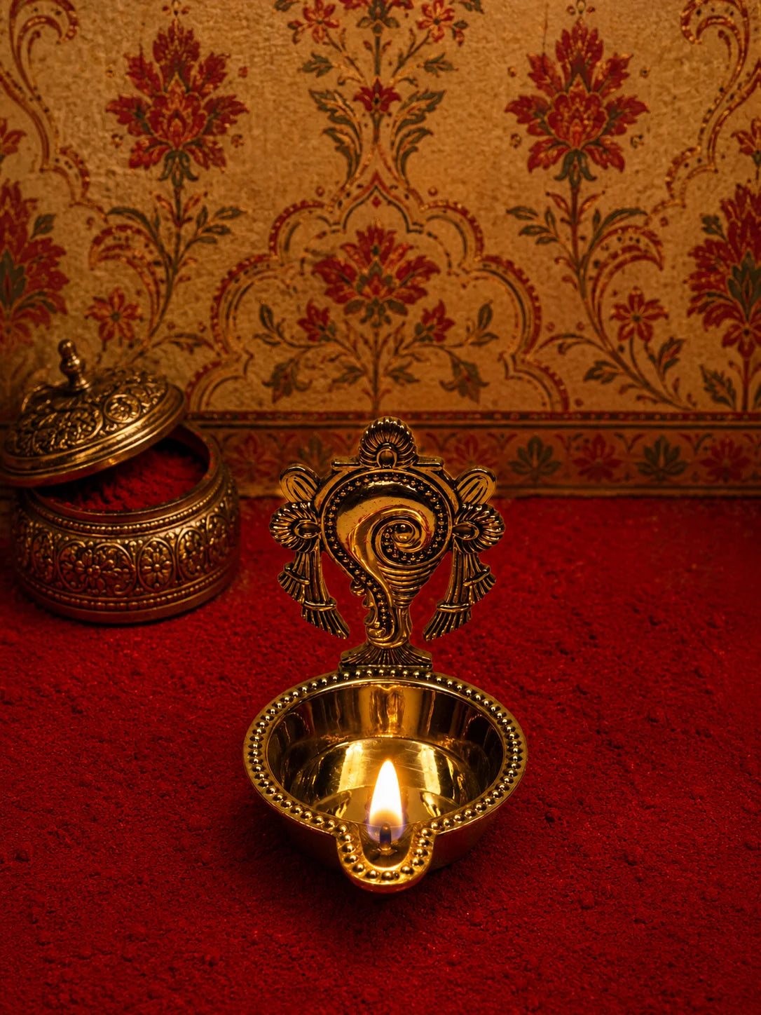 A brass diya oil lamp with a lit flame in the center, set on a red textured surface against a patterned gold and red wall, with another ornate brass vessel blurred in the background.